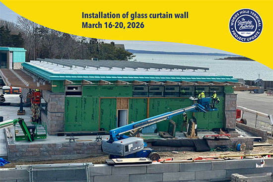 Construction workers install a glass curtain wall on a partially completed building near the water, using a blue boom lift.