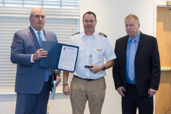 Three men stand indoors; one holds a certificate, another wears a pilot uniform holding an award, and the third wears a blue suit.