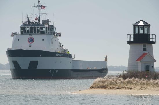 A large cargo ship named M/V Monomoy passes near a small lighthouse on a sandy shore, with calm water and clear weather.