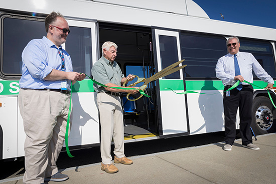 Three men stand in front of a bus; one man cuts a green ribbon with large scissors during a ribbon-cutting ceremony.