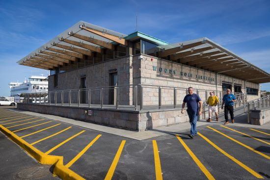 People walk outside a stone terminal building labeled Woods Hole Terminal, with a ferry visible in the background under a blue sky.